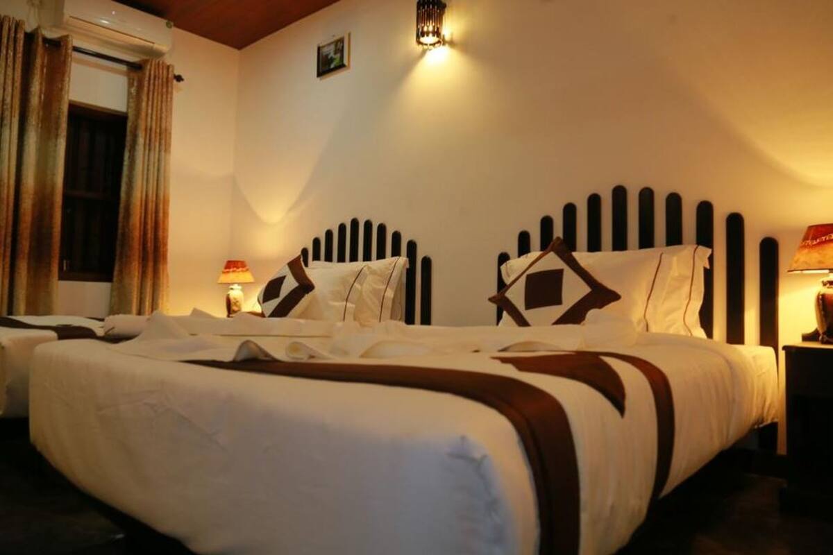 A hotel room with two beds, each with a headboard and pillows. There is a window with curtains, a wall-mounted air conditioner, and a wall-mounted light fixture.