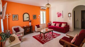 Un salon avec un canapé rouge, une table basse en bois et un tapis rouge. On y trouve une porte blanche et une fenêtre avec des cadres blancs.