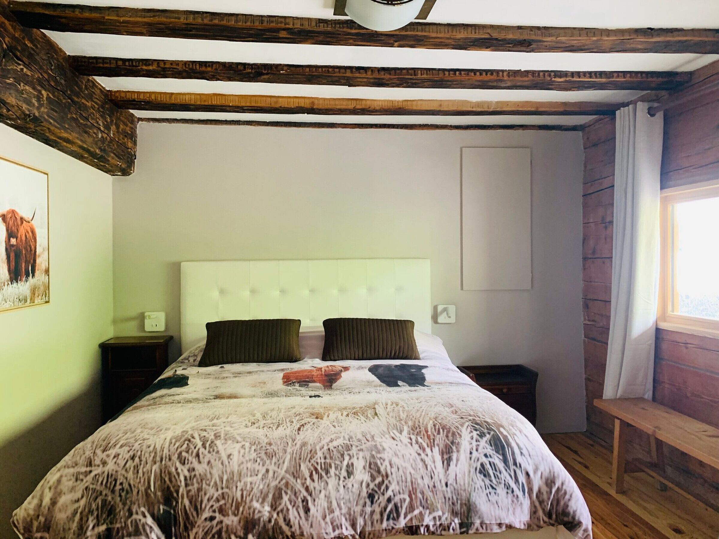 Une chambre à coucher comprenant un lit, deux tables de chevet, un banc en bois, une fenêtre avec des rideaux et un tableau accroché au mur.