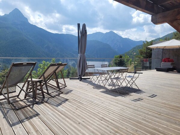 A wooden deck with outdoor furniture, a table, and a covered area, overlooking a mountainous landscape.