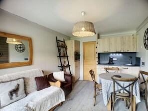 Een gezellige woonkamer met een bank, een eettafel met stoelen, een keuken met kastjes en een spoelbak, en een open wandplank.