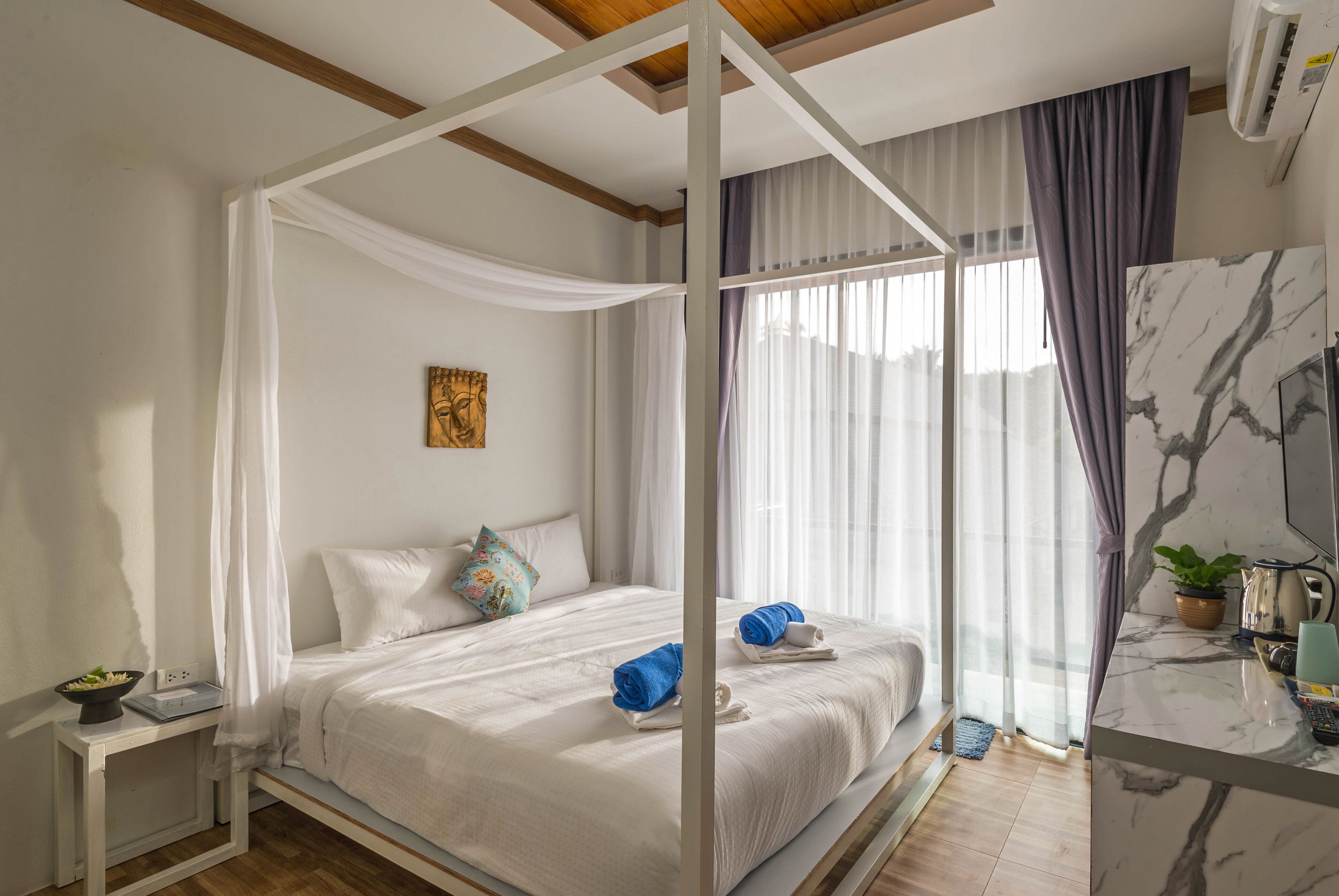 A bedroom with a four-poster bed, a bedside table with a lamp, and a view of the outdoors through large windows.