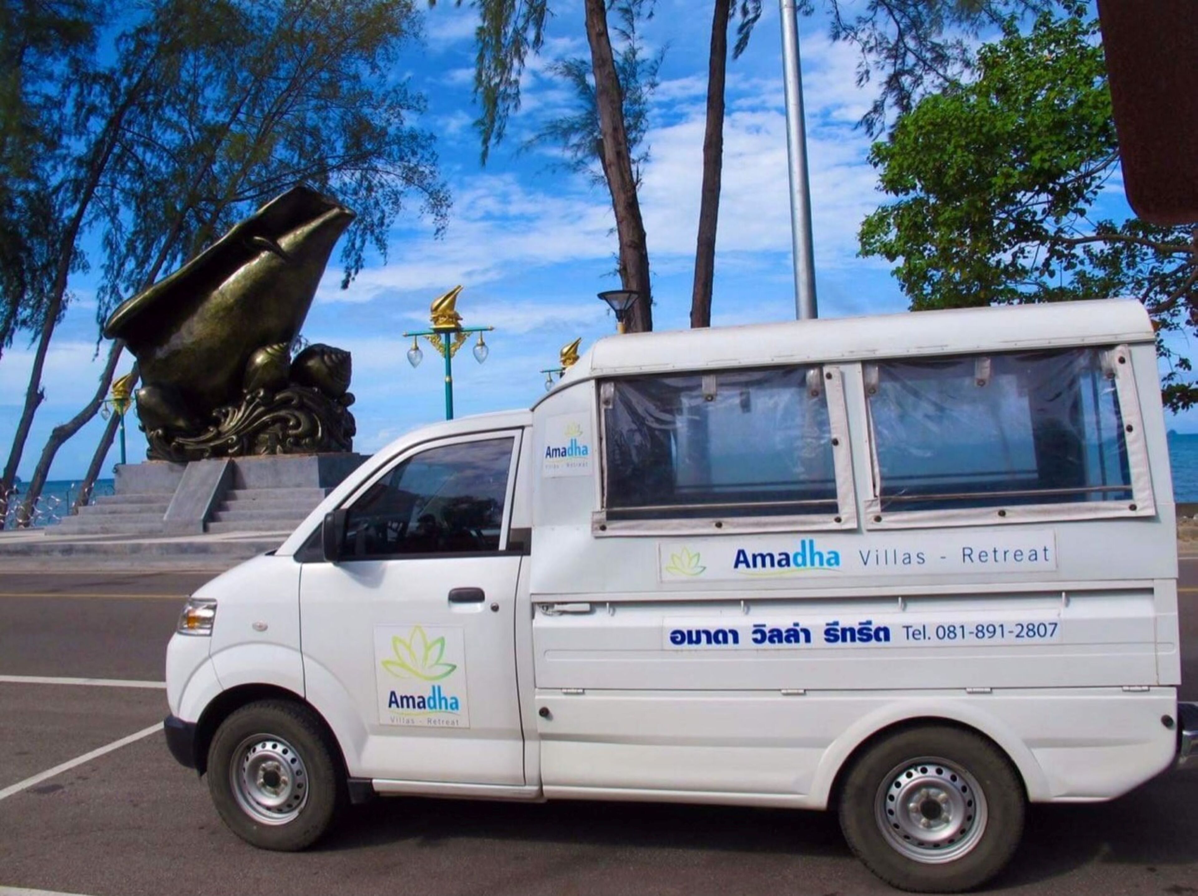 Una camioneta blanca con la marca "Amadha Villas - Retreat" estacionada frente a una estatua y árboles.