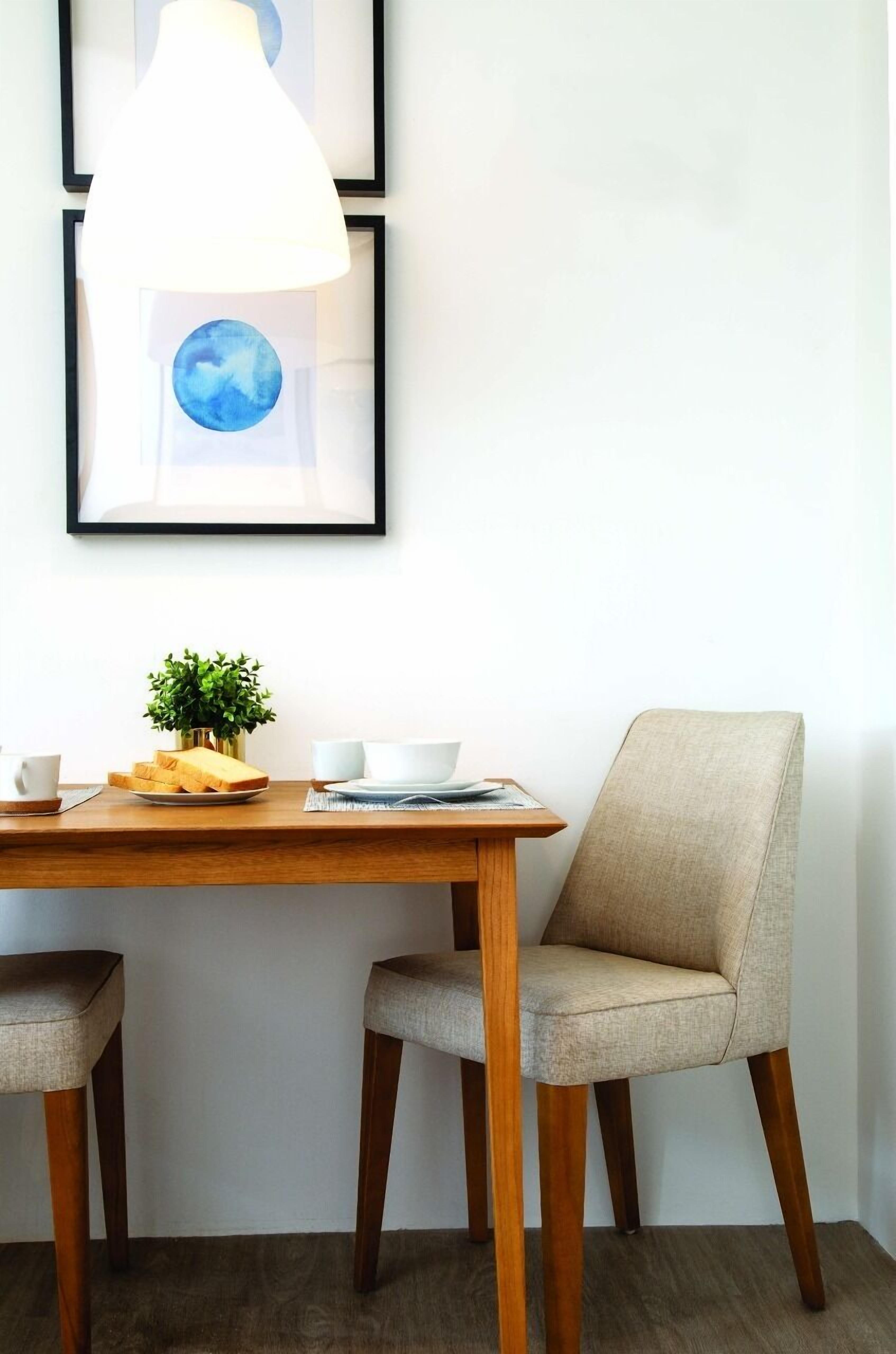 A dining area with a wooden table, two chairs, a potted plant, and framed artwork on the wall.
