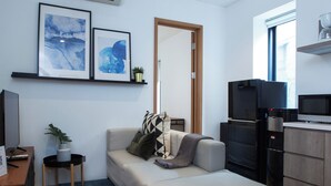A modern living room with a sofa, a TV, a kitchen area with a fridge and microwave, and artwork on the wall. - Ariama Serviced Residence (Jakarta)
