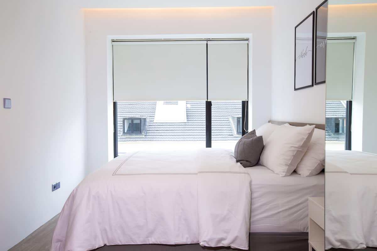 A bedroom with a large window, a bed with white bedding, and a bedside table.