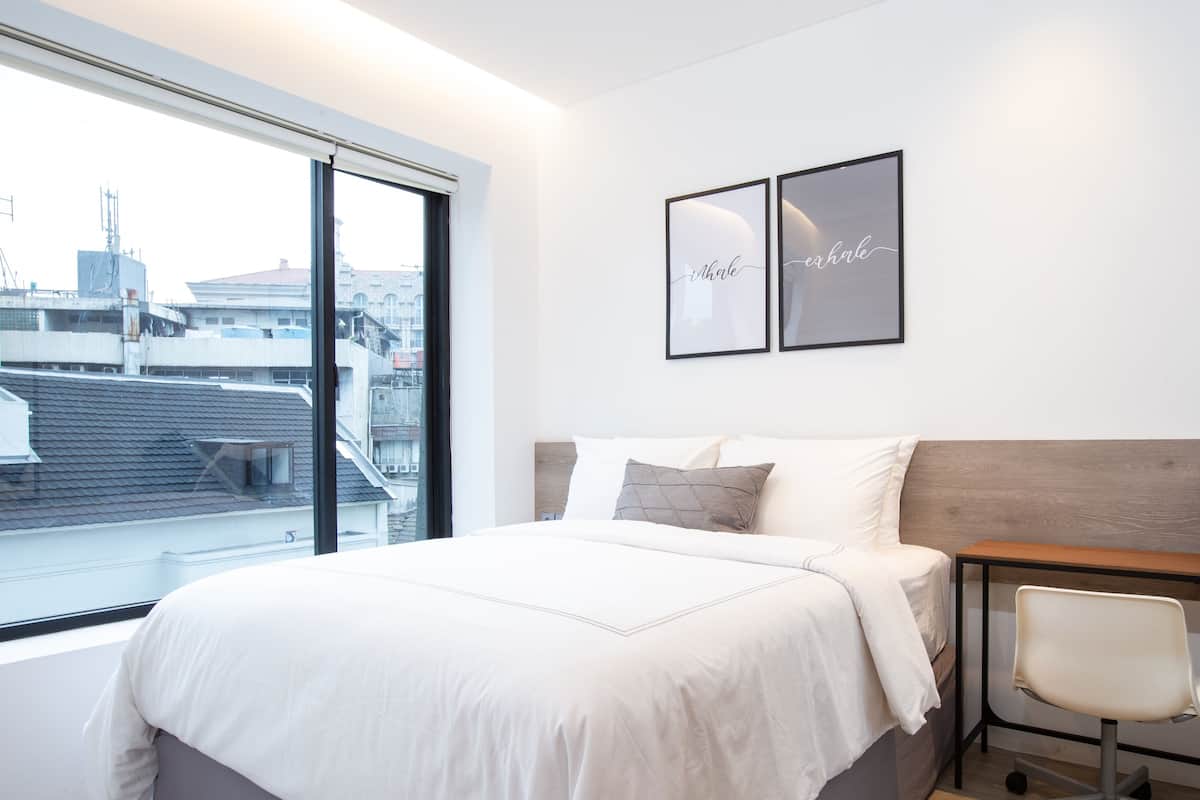 A modern hotel room with a large bed, a wooden headboard, a desk with a chair, and a city view through a large window.