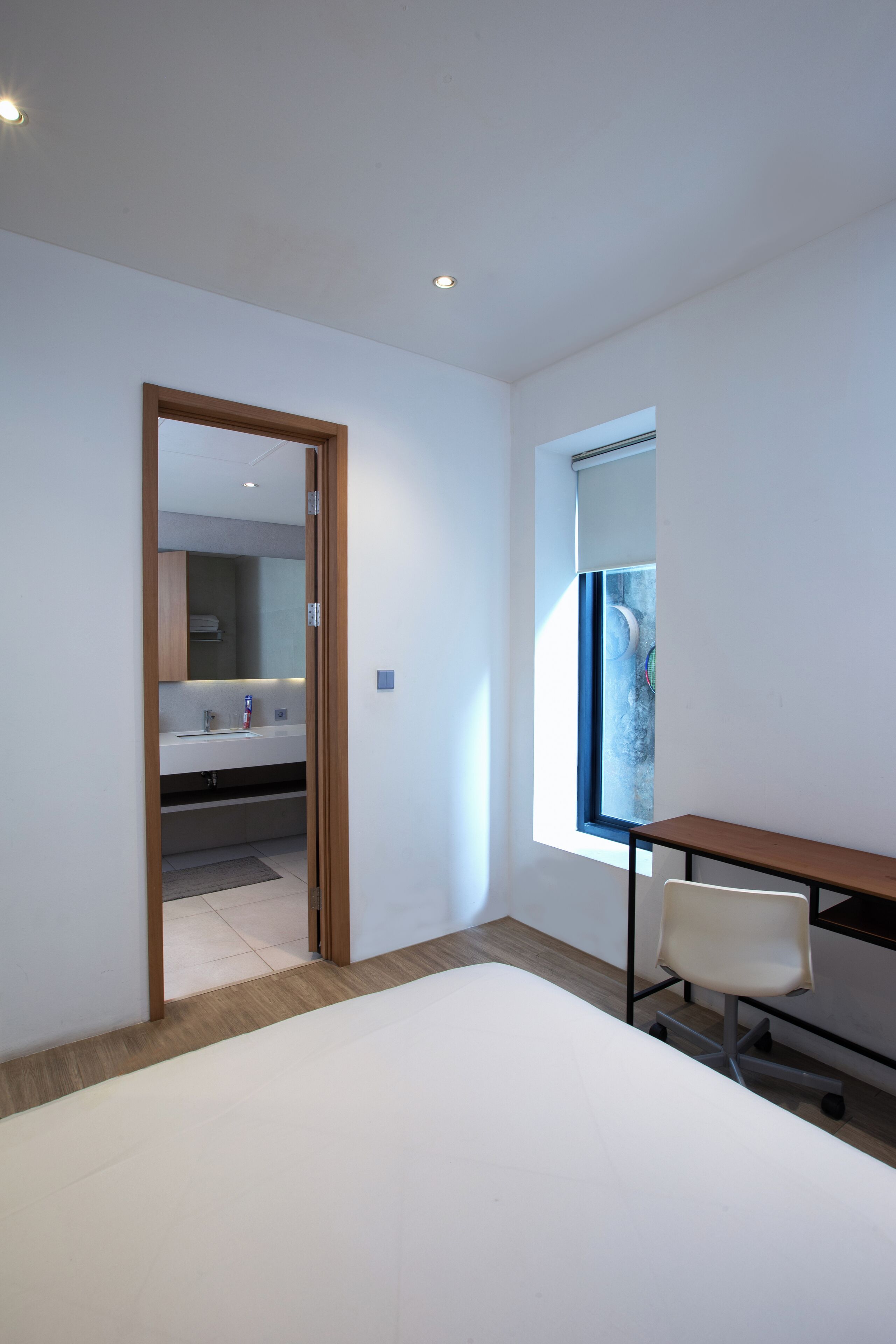 A modern bedroom with a large bed, a desk with a chair, a window with blinds, and a bathroom visible through an open door.
