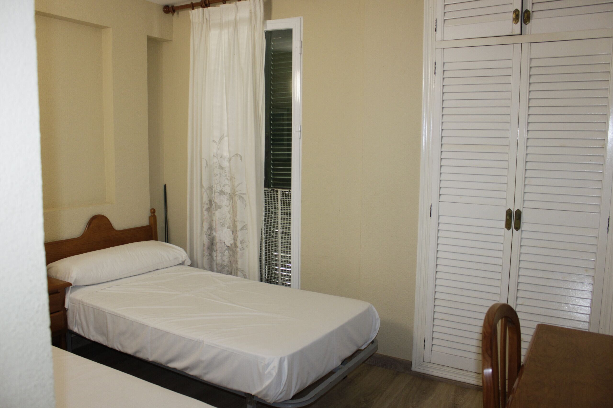 A bedroom with a single bed, a wooden nightstand, a chair, a wardrobe, and a window with curtains.