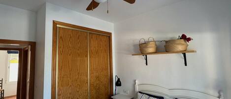 Un dormitorio con cama, un armario de madera, un ventilador de techo y dos cestas en un estante.