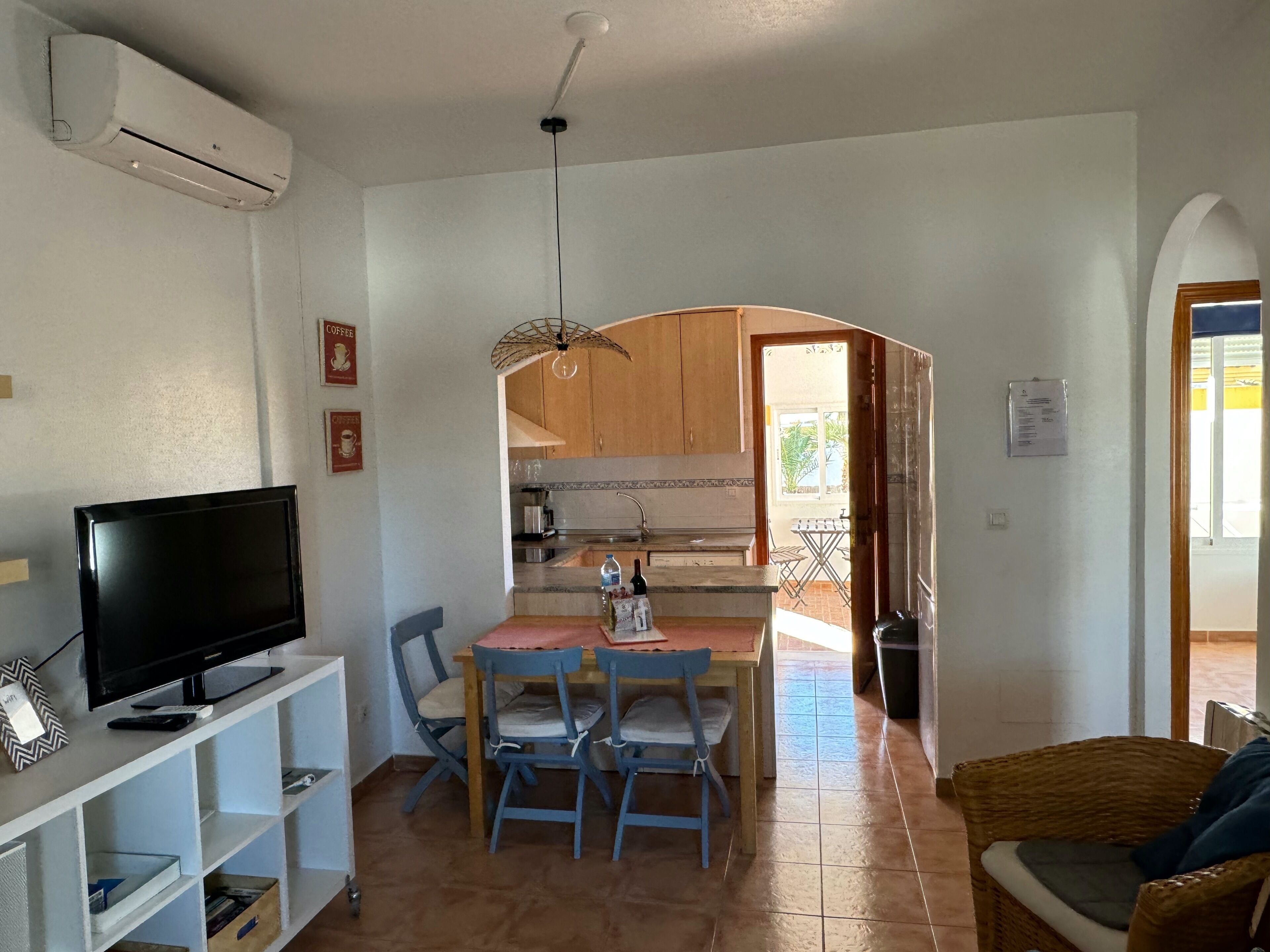 A compact living space with a TV on a shelf, a dining table with chairs, a kitchen area with wooden cabinets, and a wicker chair.