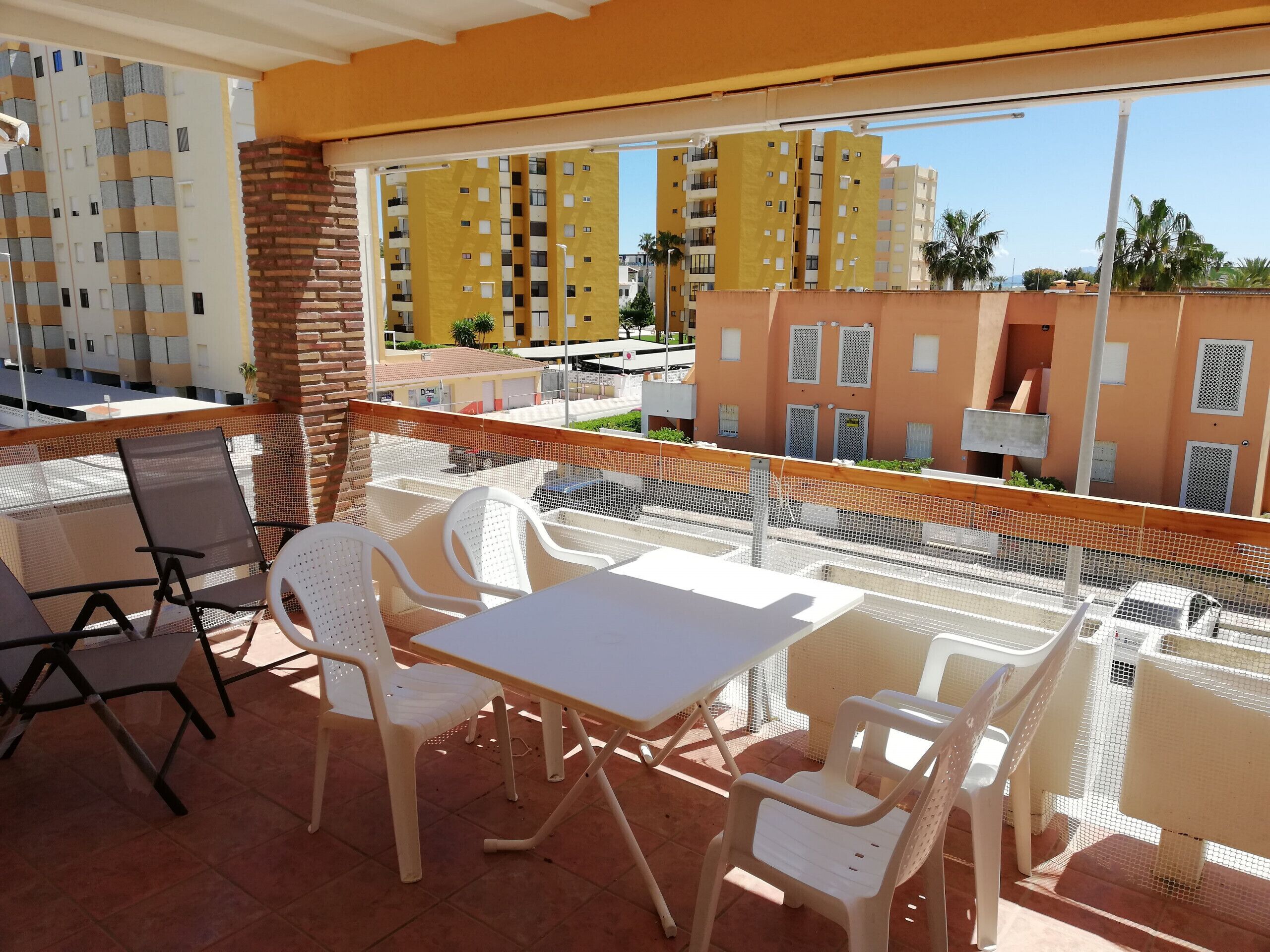 A balcony with a table and chairs, a brick pillar, and a view of yellow buildings and palm trees.