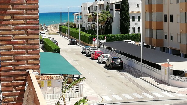 A street view with a road leading to a beach, buildings with balconies, and a clear blue sky. - Delfines Bl. VII 2º  Solo Familias! (Xeraco Playa)