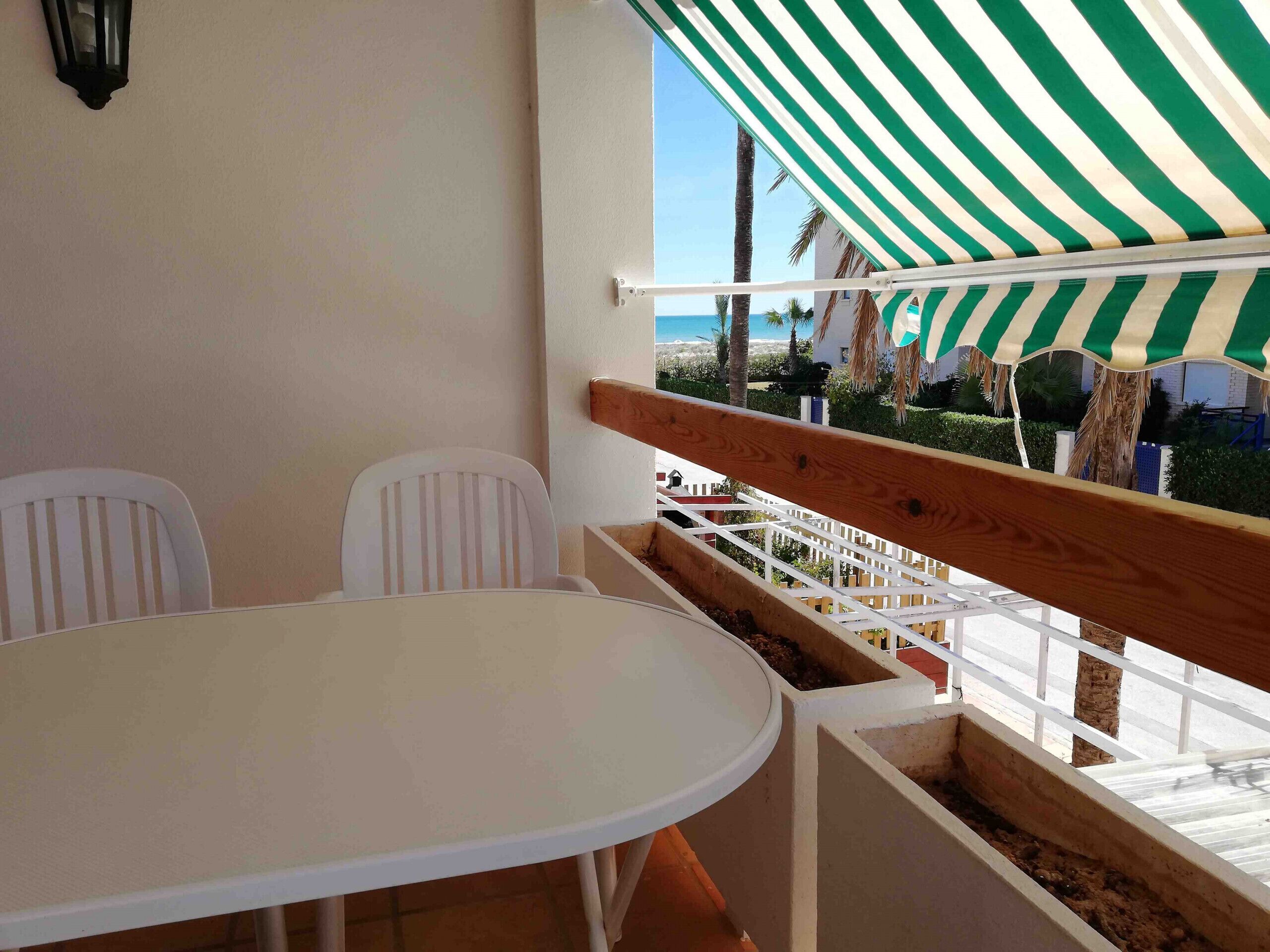 A balcony with a white table and chairs, a striped awning, and a view of palm trees and a building.