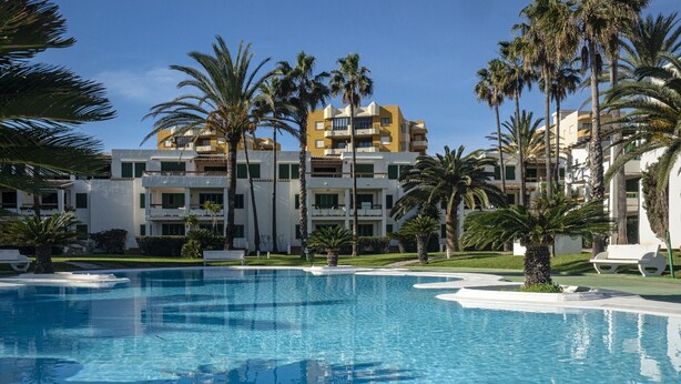 Un complexe hôtelier doté d’une piscine à eau cristalline, entourée de palmiers, et d’un immeuble à plusieurs étages avec des balcons.
