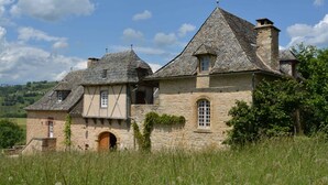 A stone building with a steeply pitched roof, a wooden door, and a window with a grid pattern. - Gite *** "La Cour pavée" (Sébrazac)