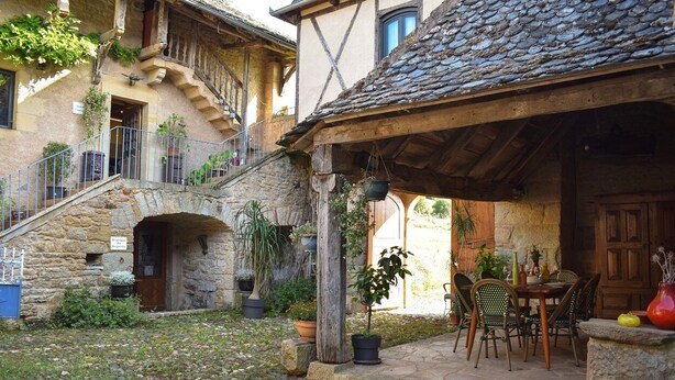 A rustic courtyard with stone flooring, a wooden table with chairs, and potted plants. - Gite *** "La Cour pavée" (Sébrazac)