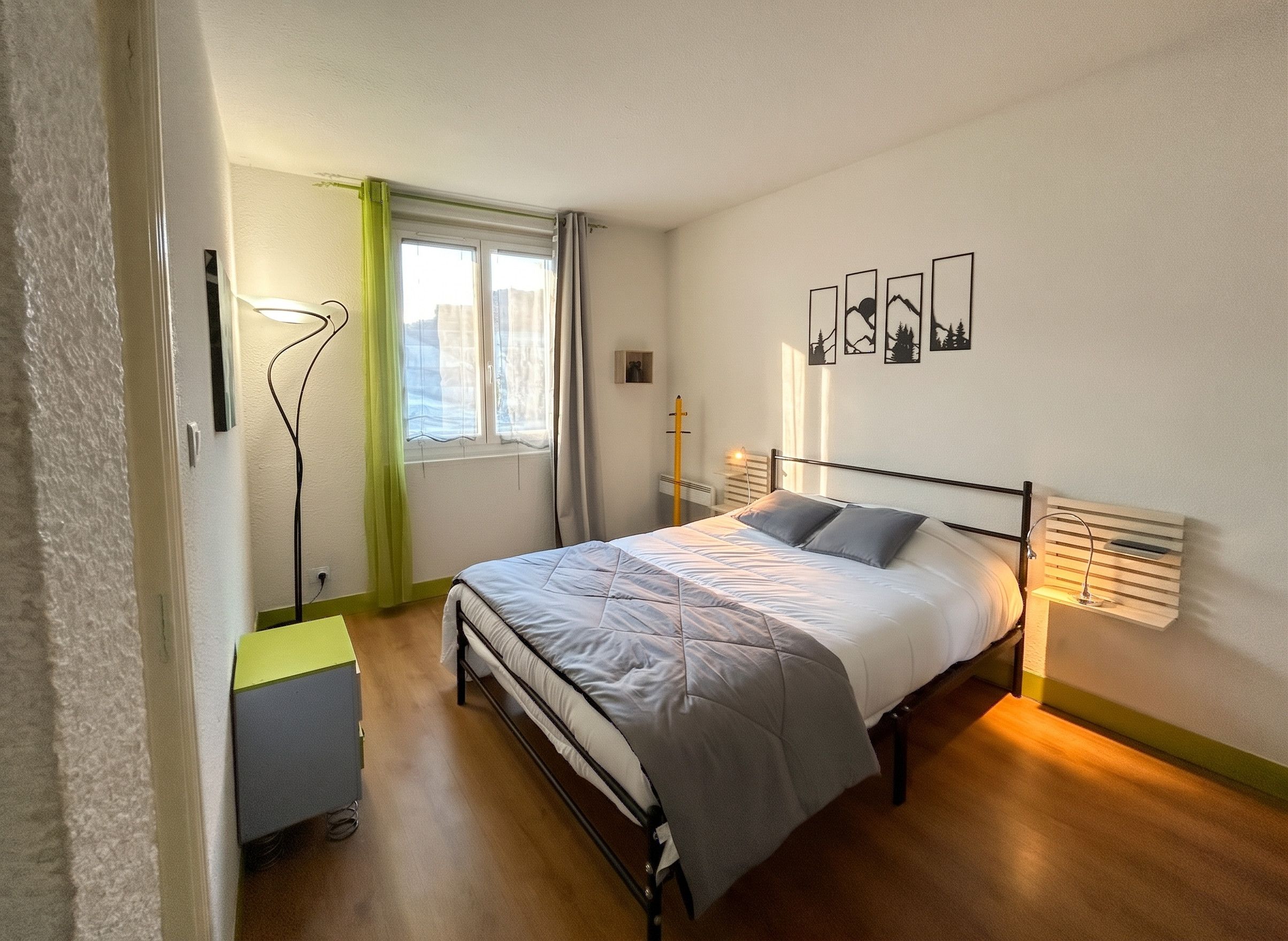 Ein Schlafzimmer mit einem Bett, einem Nachttisch, einer Lampe, einem Fenster und Wandbildern.