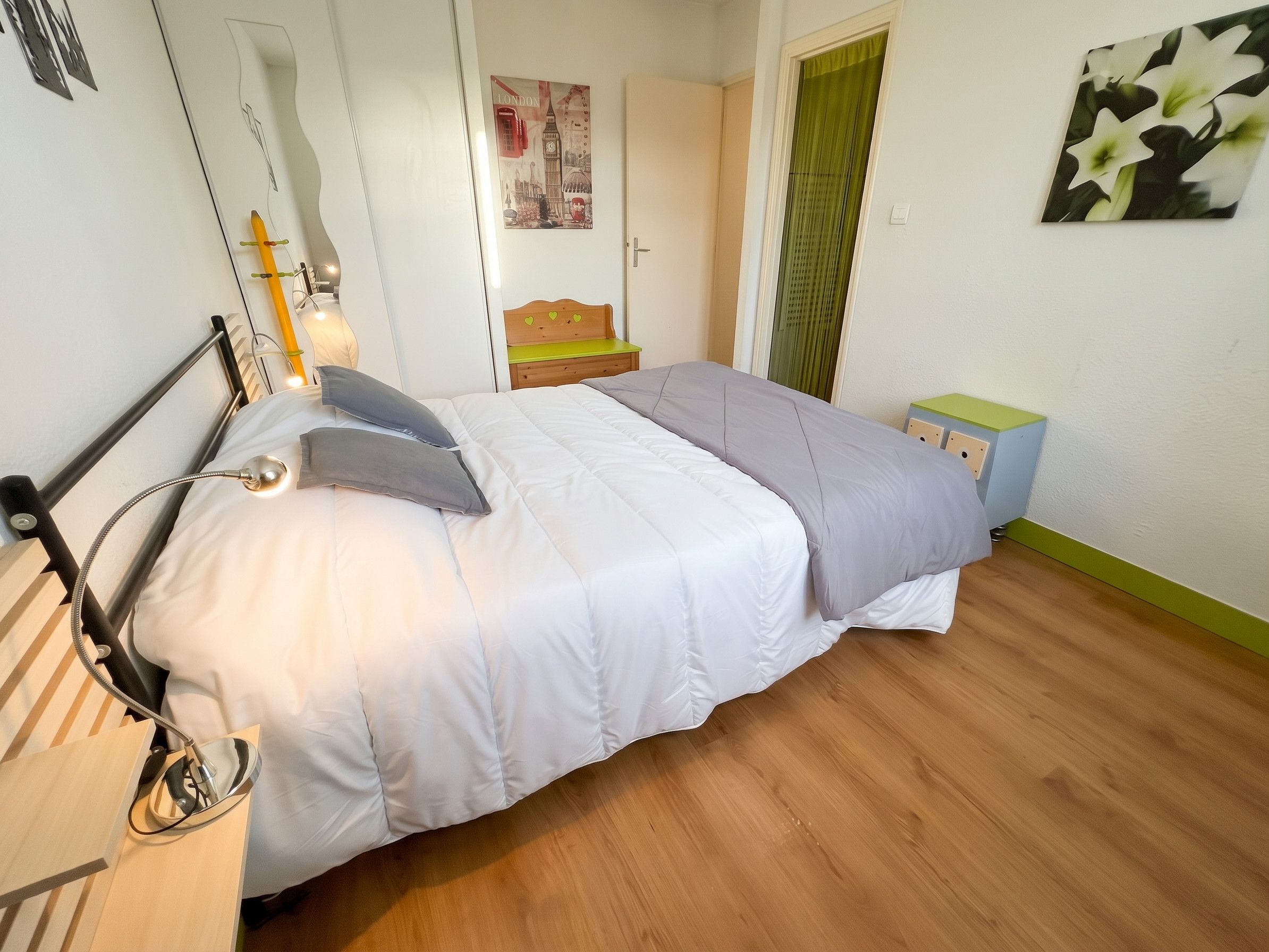 Une chambre à coucher comprenant un lit, une table de chevet, un miroir, un élément de décoration murale et une fenêtre avec des rideaux.