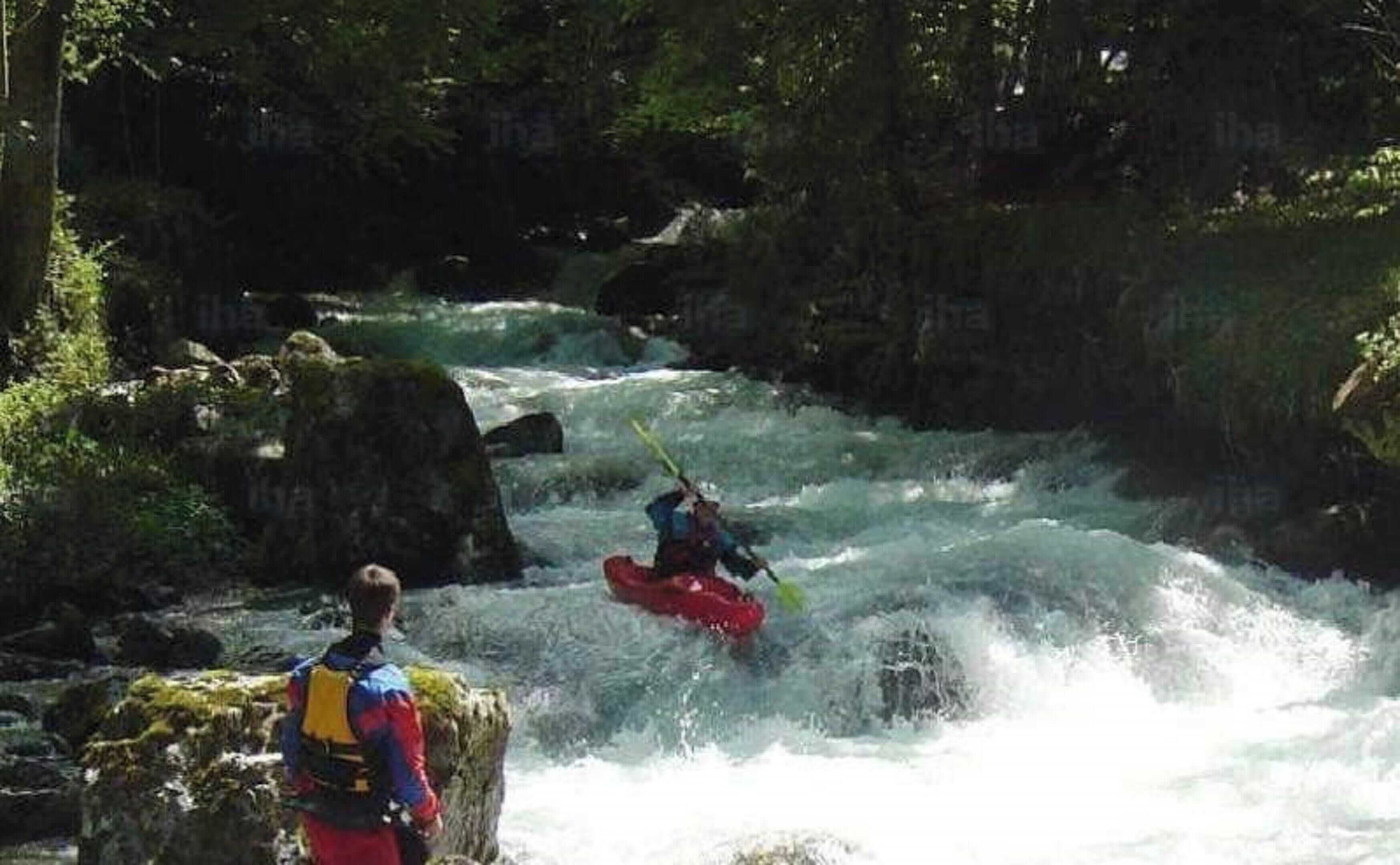 Een rivier met een persoon die kajakt en een andere persoon op rotsen staat.