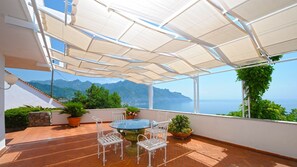 Eine überdachte Terrasse mit Tisch, Stühlen, Kübelpflanzen und Blick auf Meer und Berge.