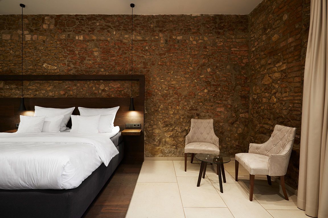 Un dormitorio con pared de piedra, una cama con ropa blanca, dos sillones y una pequeña mesa.