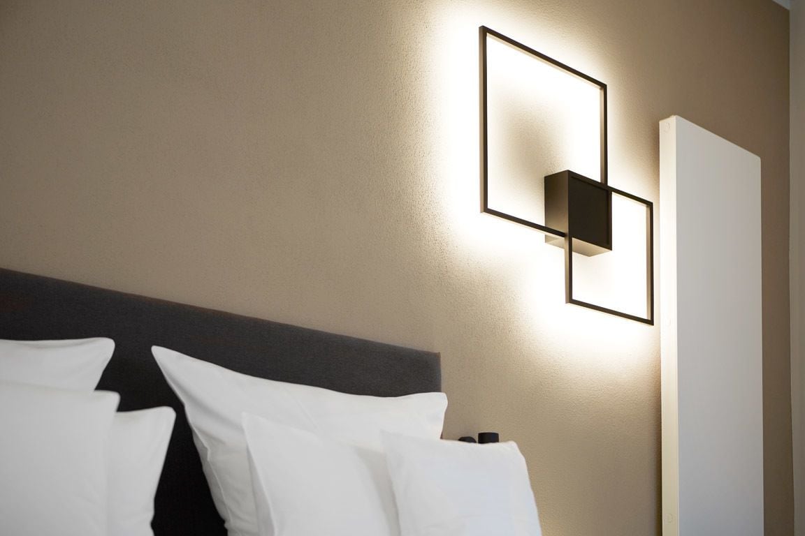 A hotel room with a modern headboard, a wall-mounted light fixture, and a large window.