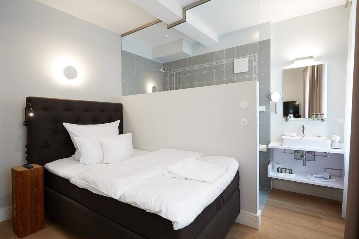 A modern bedroom with a large bed, a bedside table, a built-in shower, and a bathroom with a sink and mirror.