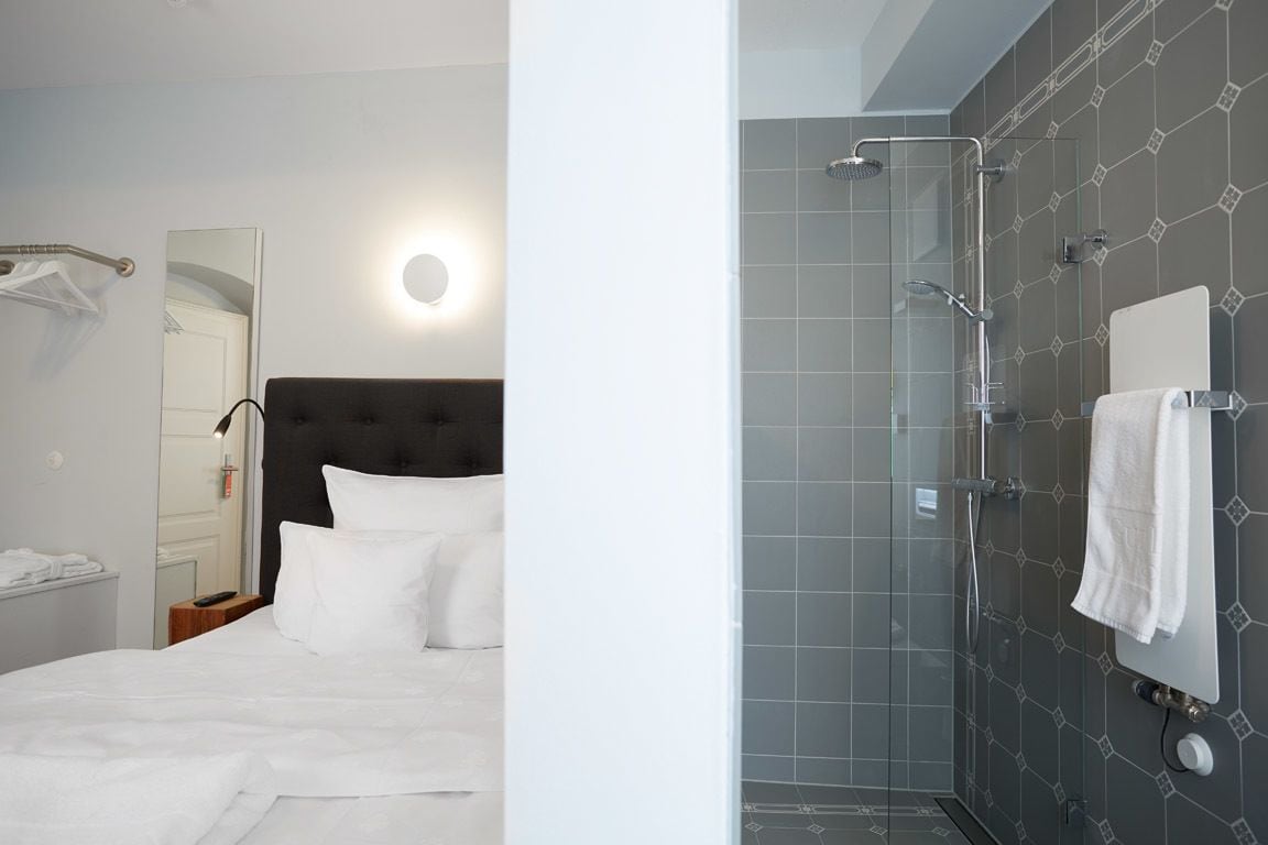 Quarto de hotel com cama, mesa de cabeceira, espelho, área de chuveiro com porta de vidro e porta-toalhas.