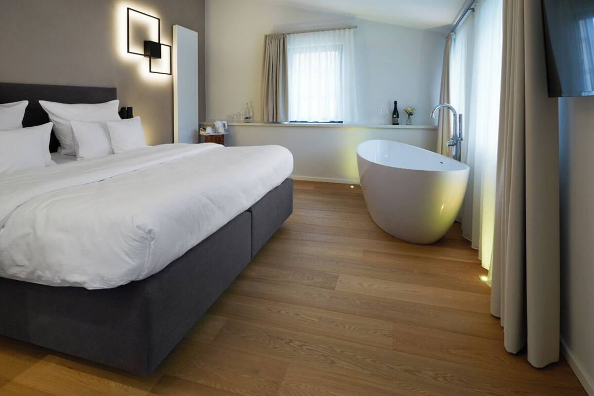 A modern hotel room with a large bed, a bathtub, wooden flooring, and a window with curtains.