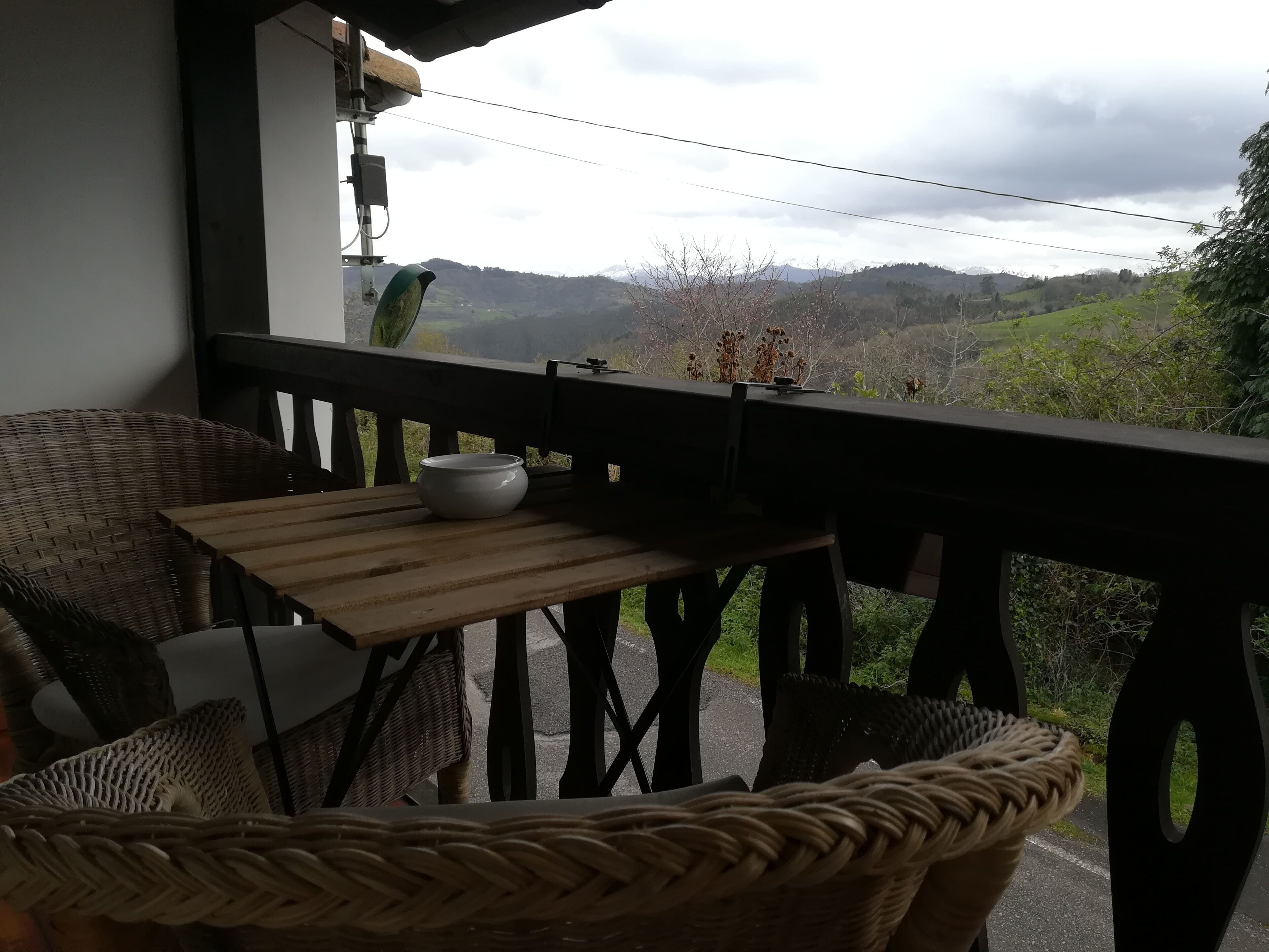 Un balcó amb mobles de vusta, una taula i vista a un paisatge amb muntanyes.