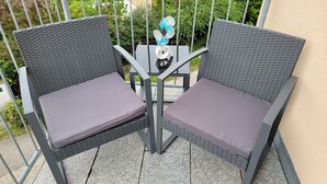 Zwei Korbstühle mit Kissen auf einem Balkon mit Metallgeländer und einem kleinen Tisch mit einer Dekoration.