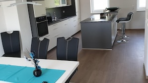 Eine moderne Küche mit weißen Schränken, einer schwarzen Arbeitsplatte und einem eingebauten Ofen. Ein Essbereich mit einem weißen Tisch, schwarzen Stühlen und einer blauen Tischmatte.