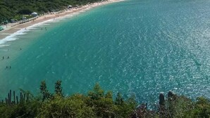 A coastal landscape with a beach, clear turquoise waters, and dense green hills in the background. - Pousada Roça Velha (Arraial do Cabo)