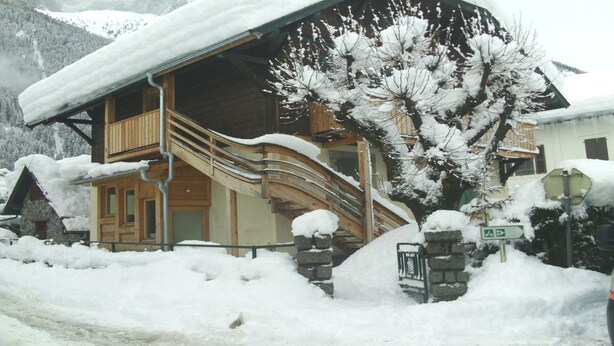 Een houten chalet met een houten trap naar een balkon, omringd door met sneeuw bedekte bomen en een berg op de achtergrond.