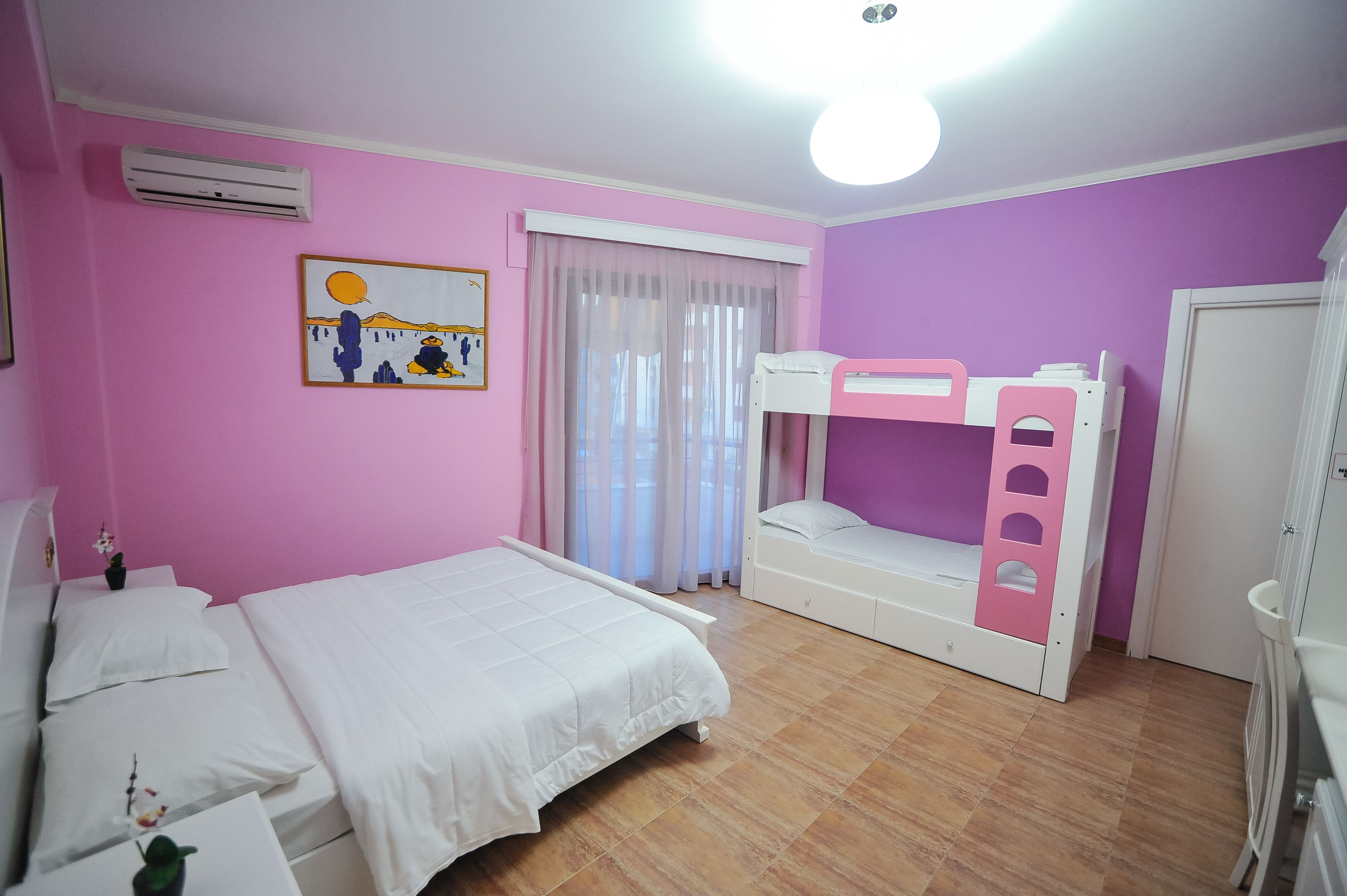 A bedroom with a bunk bed, a single bed, a window with sheer curtains, and a wall-mounted air conditioner.