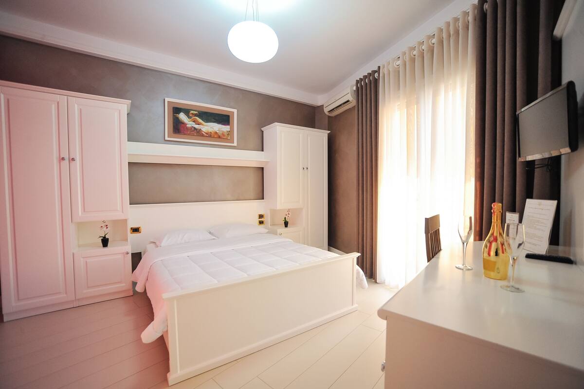 A hotel room with a bed, a desk with a bottle and glasses, a TV, and a painting on the wall.