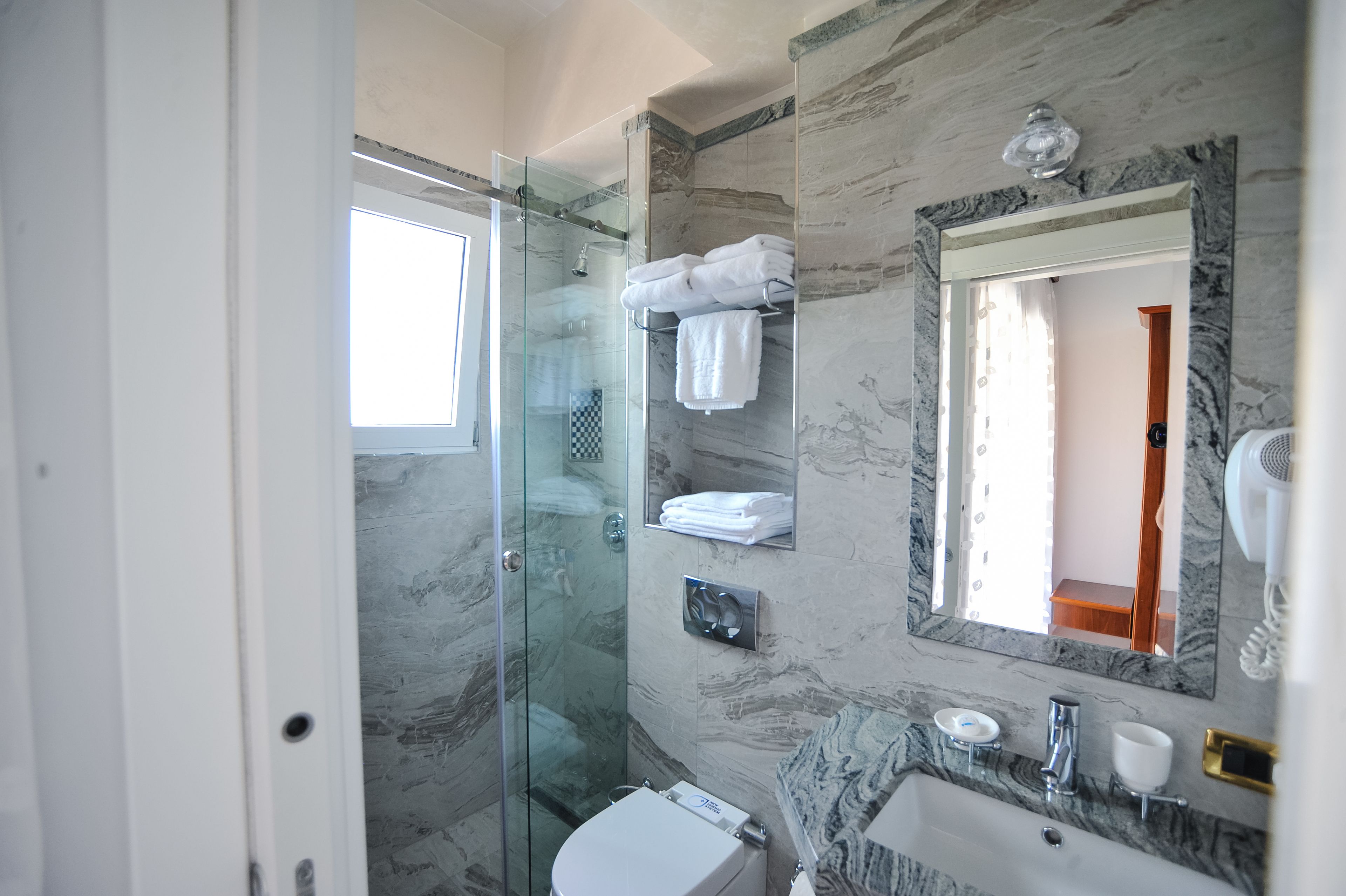 A modern bathroom with a glass shower enclosure, marble walls, a large mirror, and a white toilet.