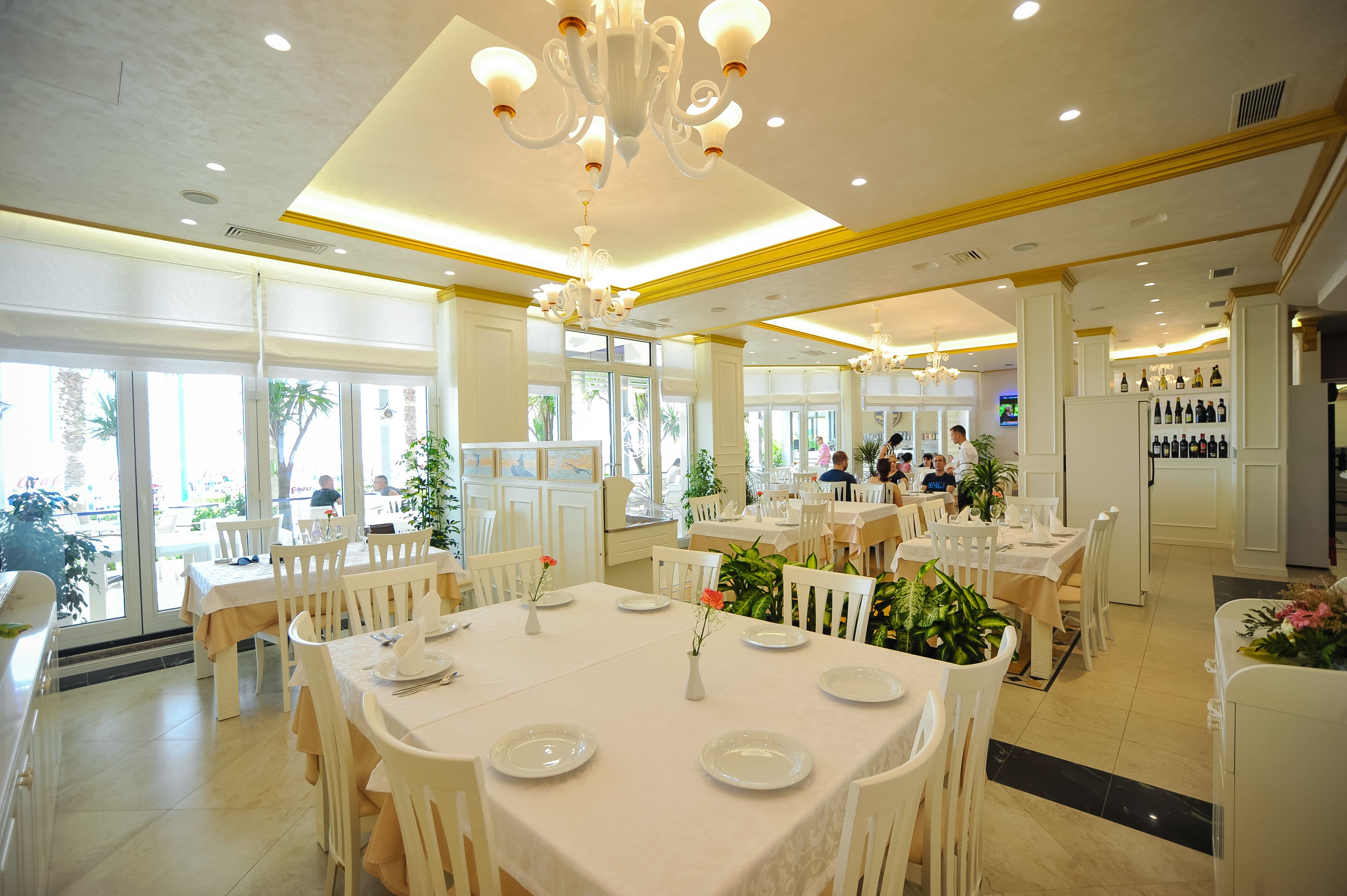 A spacious restaurant with white tables and chairs, large windows, and a chandelier.