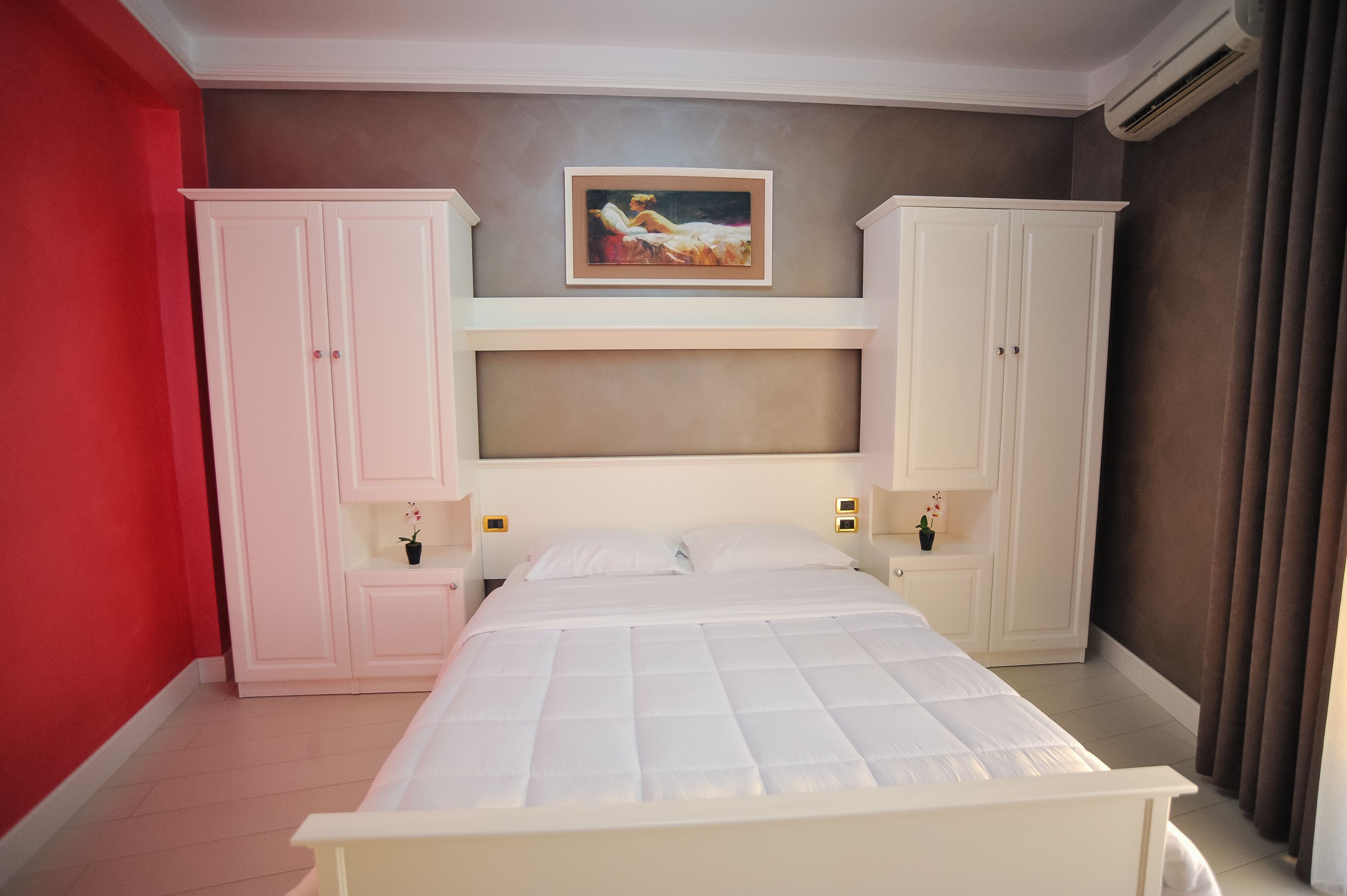 Une chambre à coucher avec un lit, deux armoires blanches, un tableau au-dessus du lit et un mur rouge sur un des côtés.