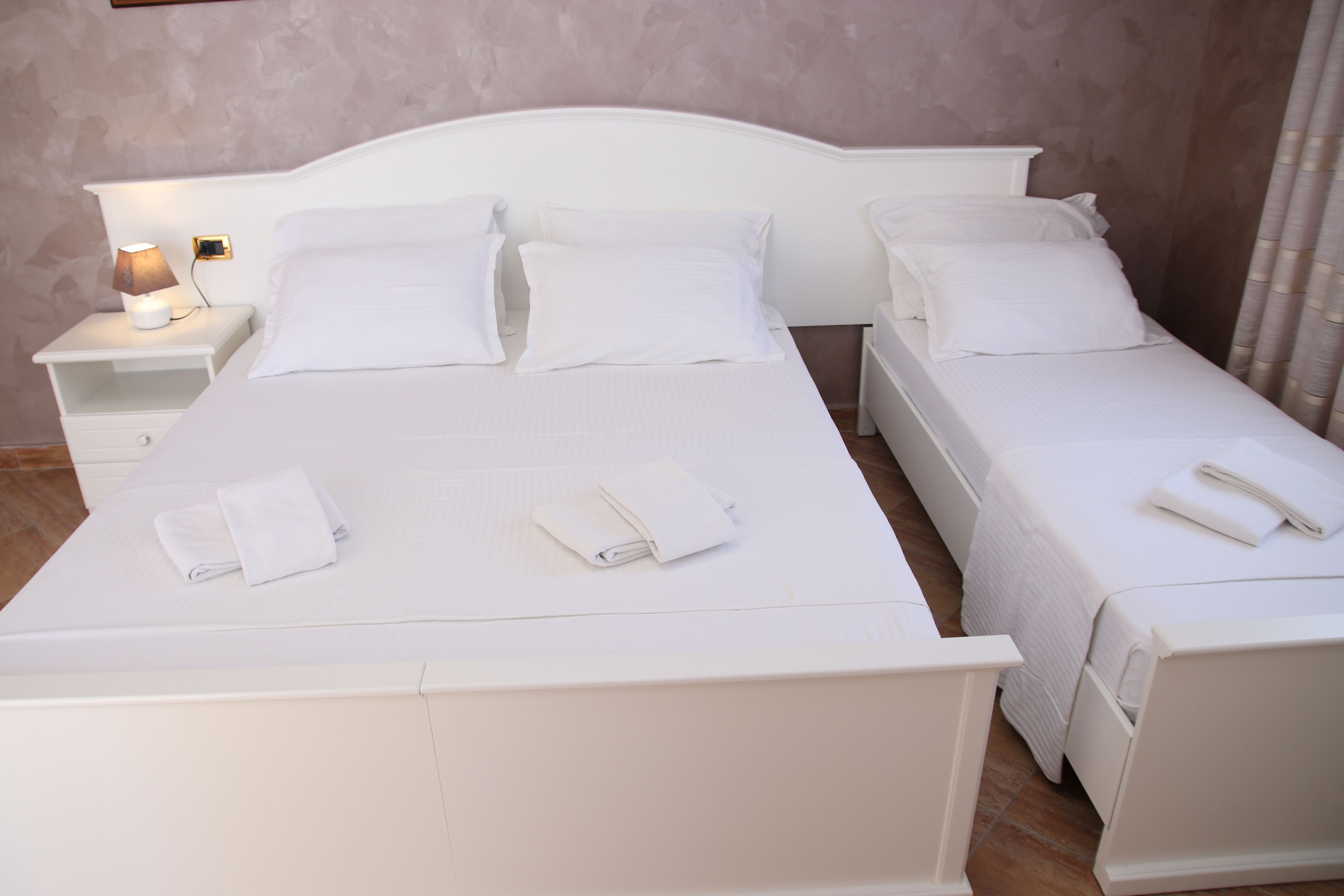 Cama de casal com lençóis e almofadas brancas, mesa de cabeceira com abajur e criado-mudo com telefone.
