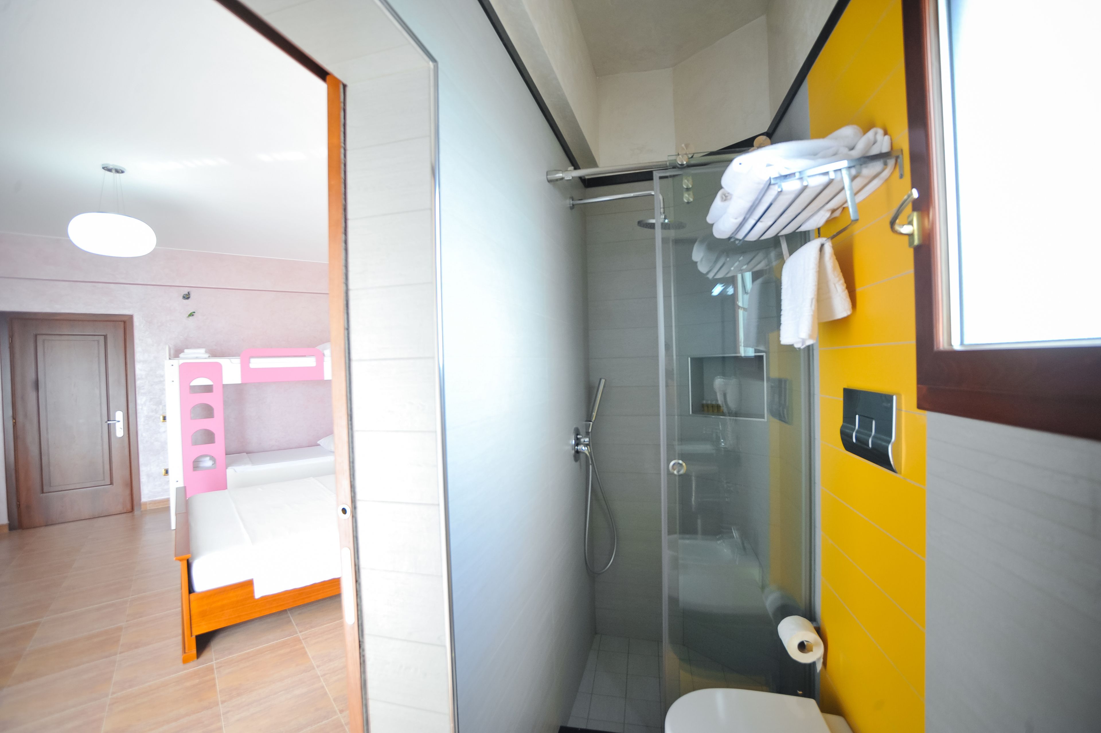A modern bathroom with a glass shower enclosure, a toilet, and a bed in the adjacent room.