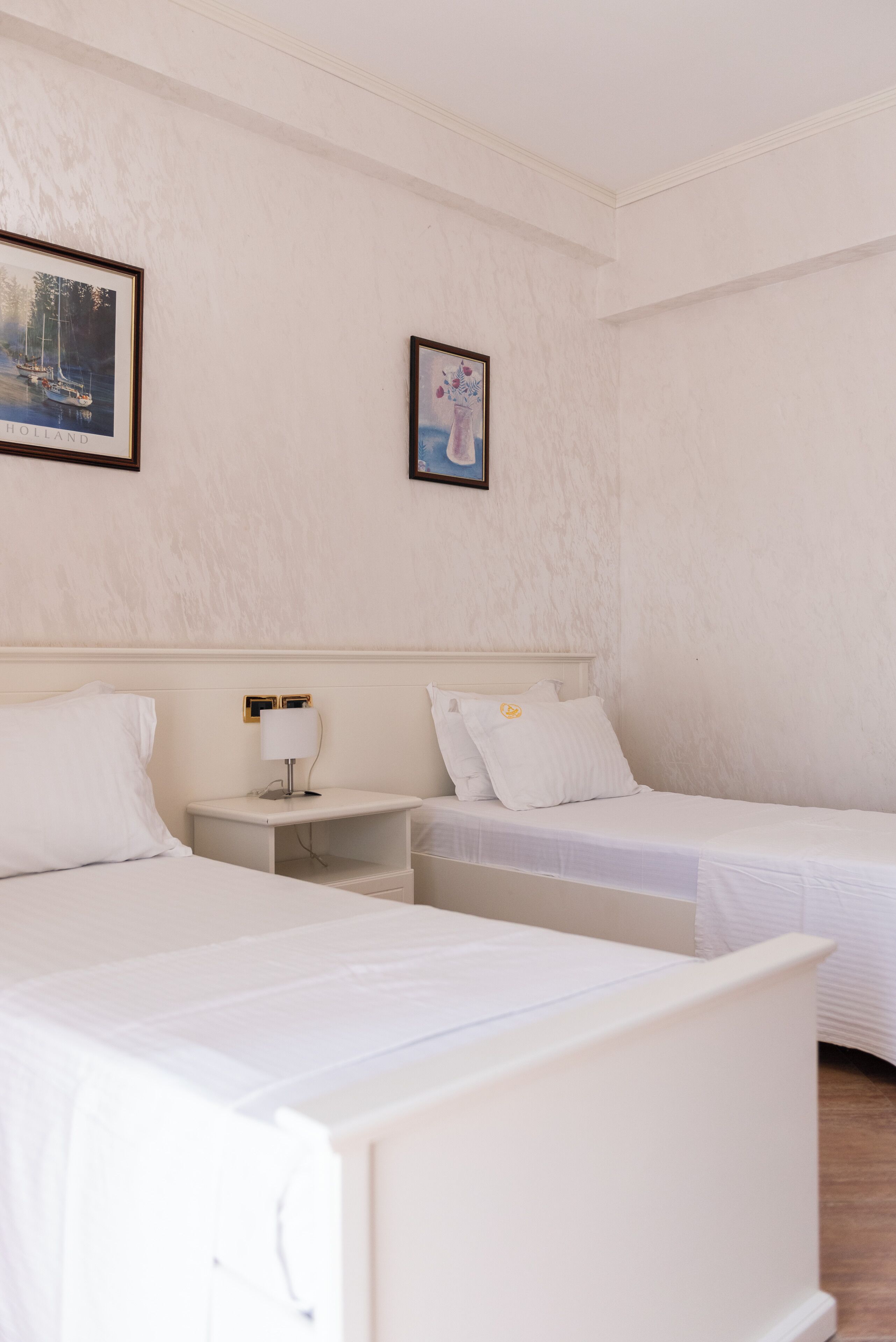 A hotel room with two single beds, a nightstand with a lamp, and two framed pictures on the wall.