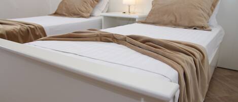 A hotel room with two single beds, each with a white mattress and a beige blanket. There is a bedside table with a lamp between the beds.