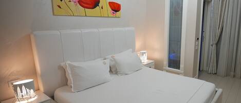 A bedroom with a large bed, white bedding, a nightstand with a lamp, and a painting of red flowers on the wall.