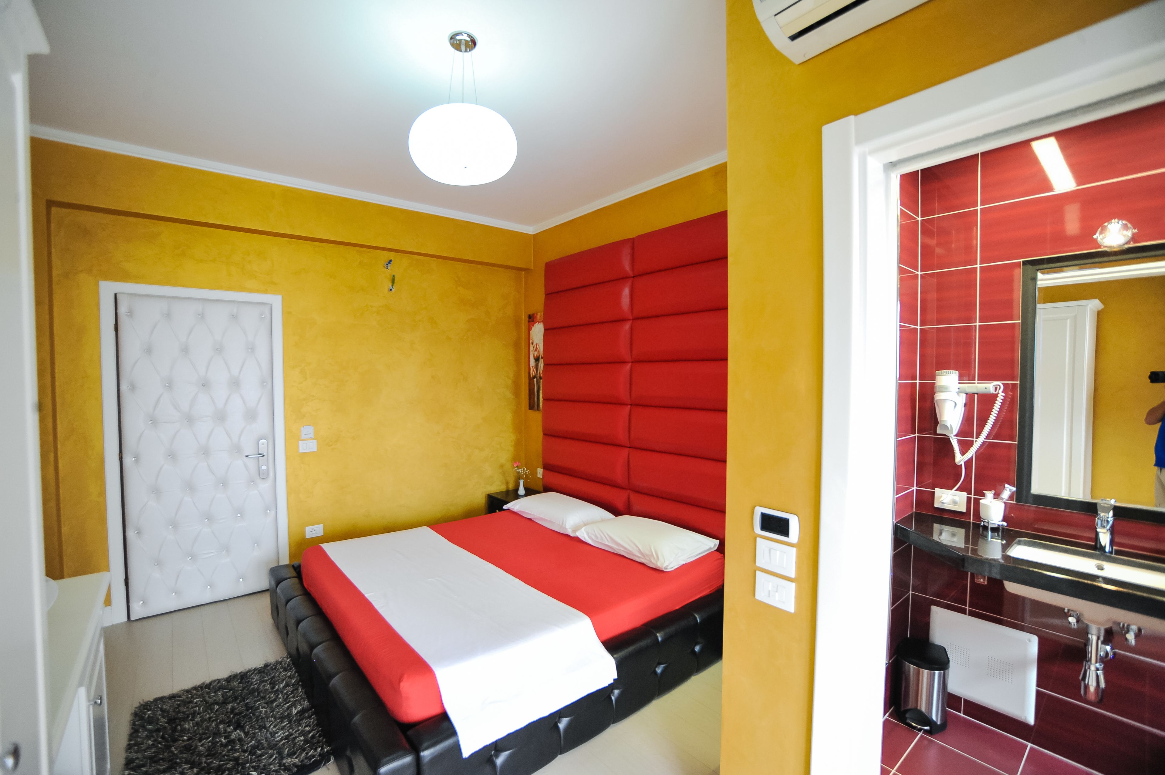Une chambre d’hôtel avec un lit, une porte blanche, une tête de lit rouge, une salle de bain avec un mur recouvert de carreaux rouges et un lavabo.