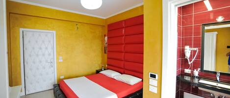 Une chambre d’hôtel avec un lit, une porte blanche, une tête de lit rouge, une salle de bain avec un mur recouvert de carreaux rouges et un lavabo.
