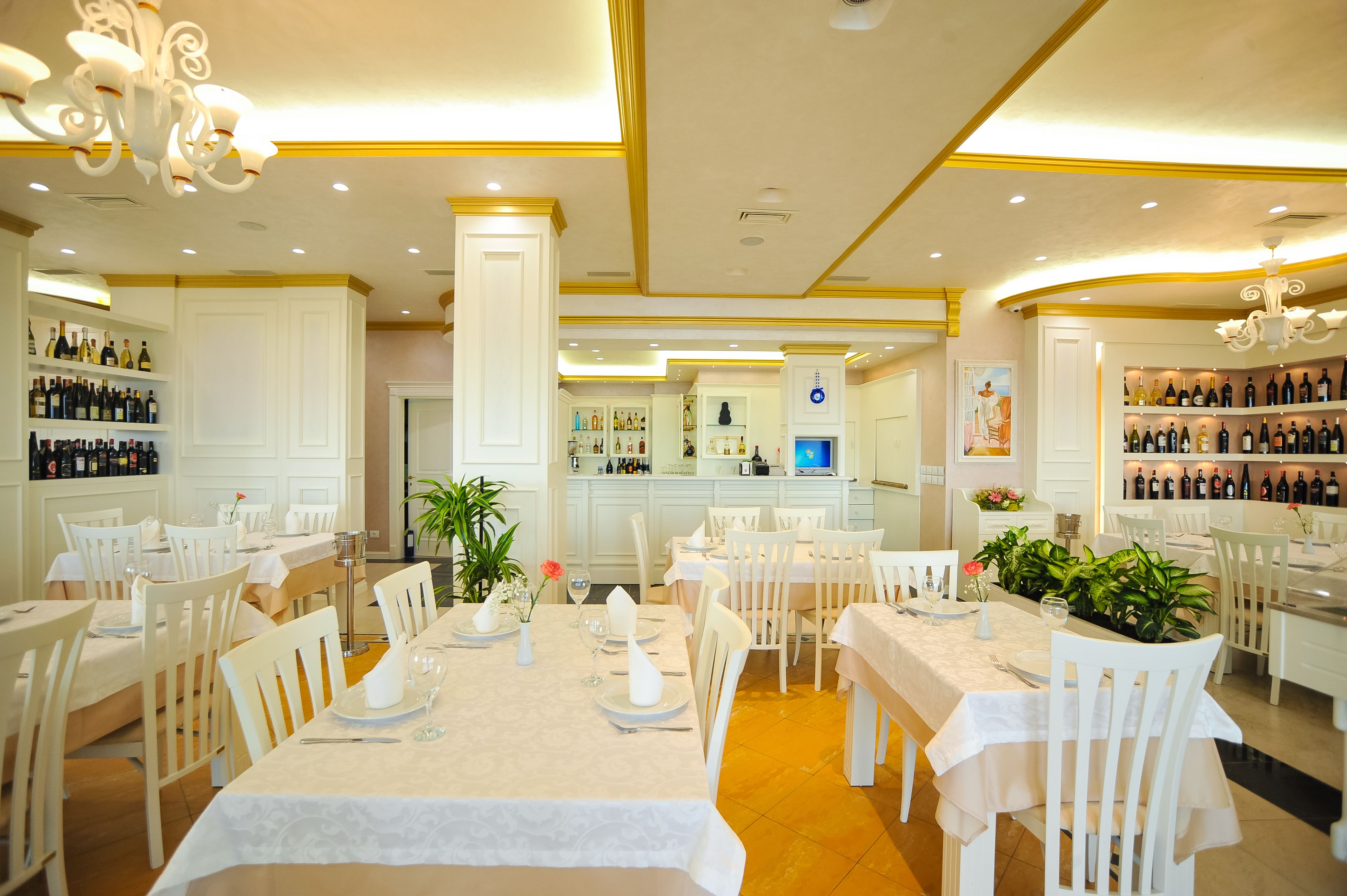 Un ristorante con tavoli e sedie bianche, un'area bar con scaffali pieni di bottiglie e un pilastro centrale.