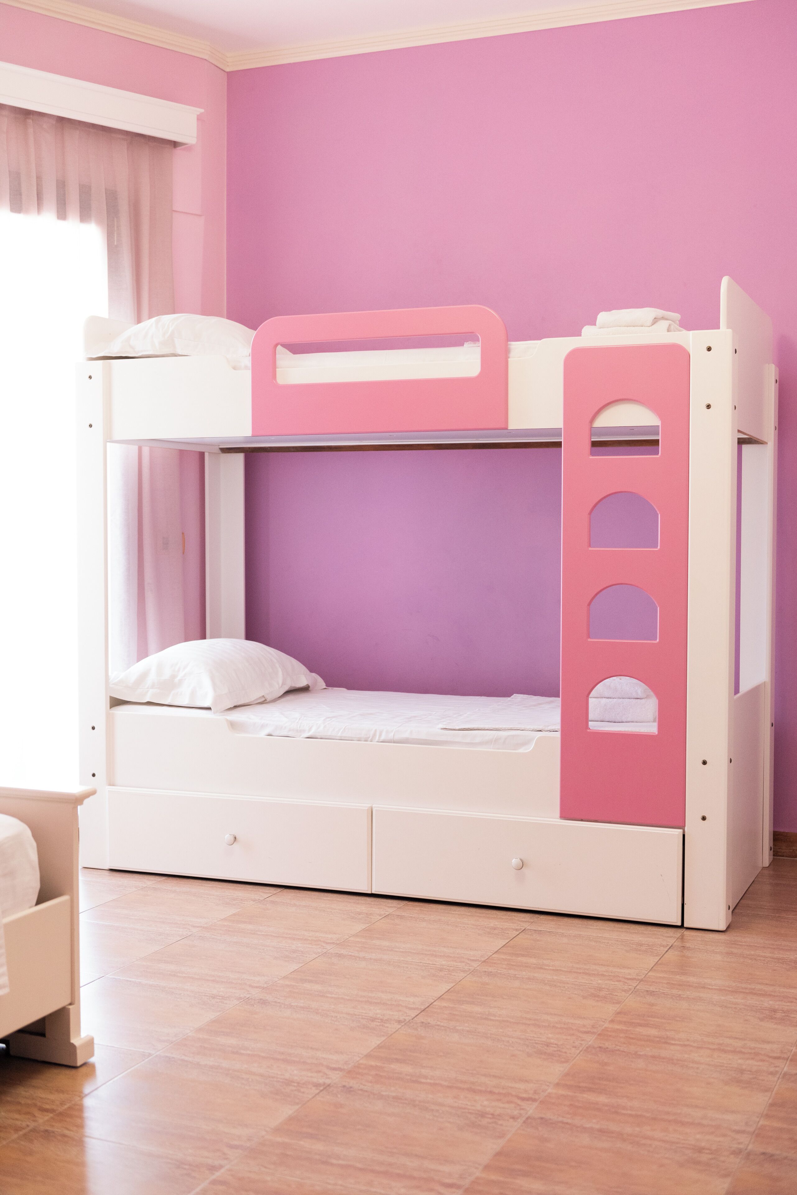 Una litera con escalera rosa y una parte inferior blanca en una habitación con paredes moradas y una ventana con cortinas.