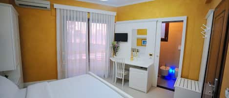 Quarto de hotel com cama, escrivaninha com cadeira, televisão e banheiro visível por uma porta aberta.